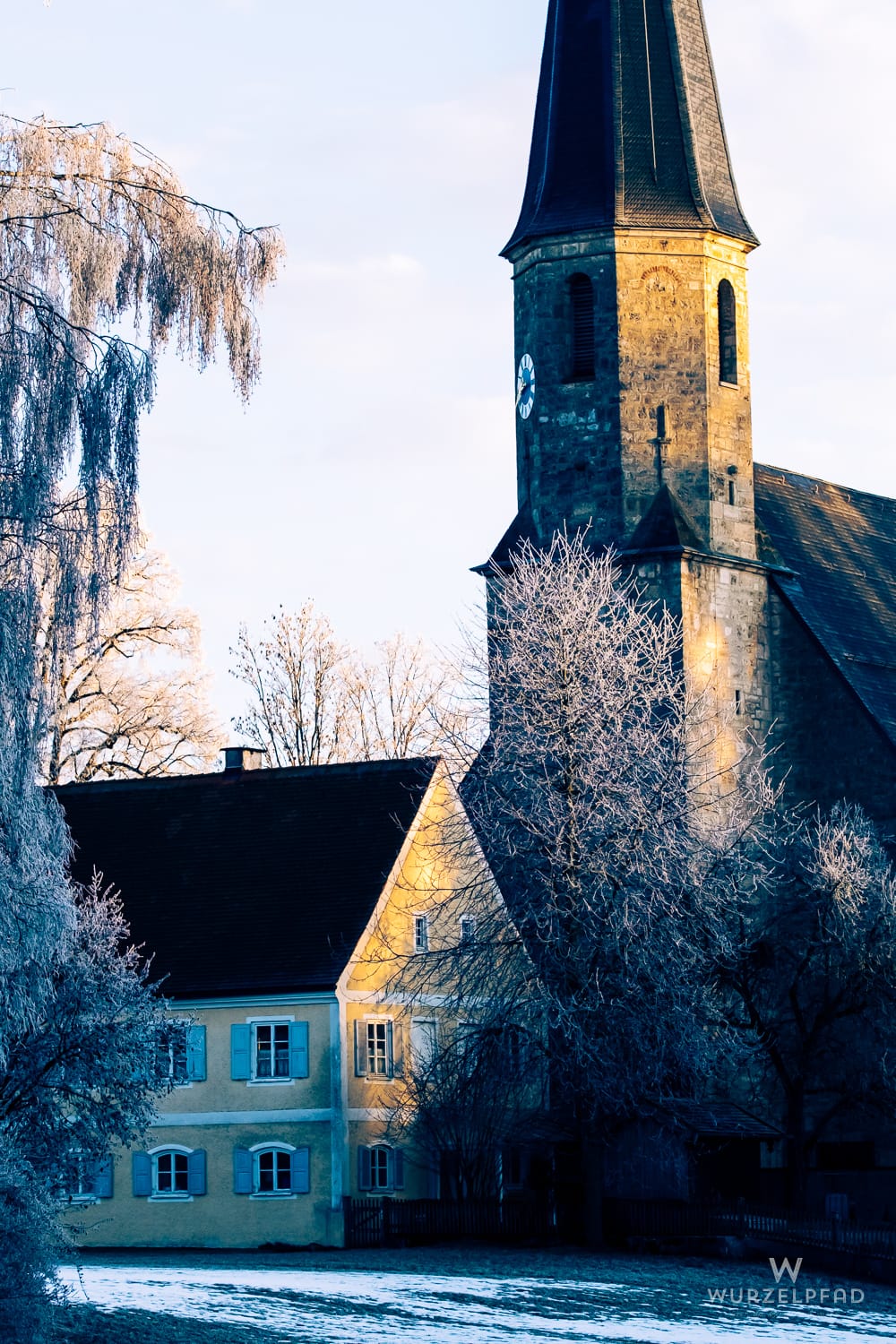Kirche St. Johann Baptist in Föching: Steinerner Turm und gelbes Haus im Sonnenlicht, umgeben von raureifbedeckten Bäumen und Schnee. Winterliche Szene.