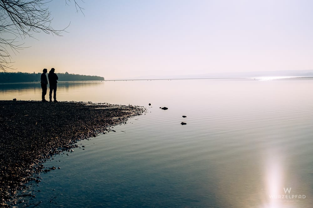 Zwei Spaziergänger stehen als Silhouette am steinigen Seeufer und blicken über das ruhige Wasser, das das helle Licht des Himmels spiegelt. Friedliche Stimmung.