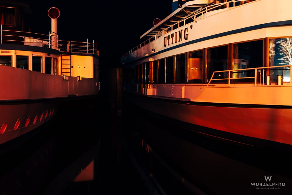 Zwei Ammerseedampfer, darunter die 'UTTING', liegen nachts nebeneinander im Hafen, künstlich beleuchtet in warmen Orange- und Rottönen, mit Spiegelungen im Wasser.