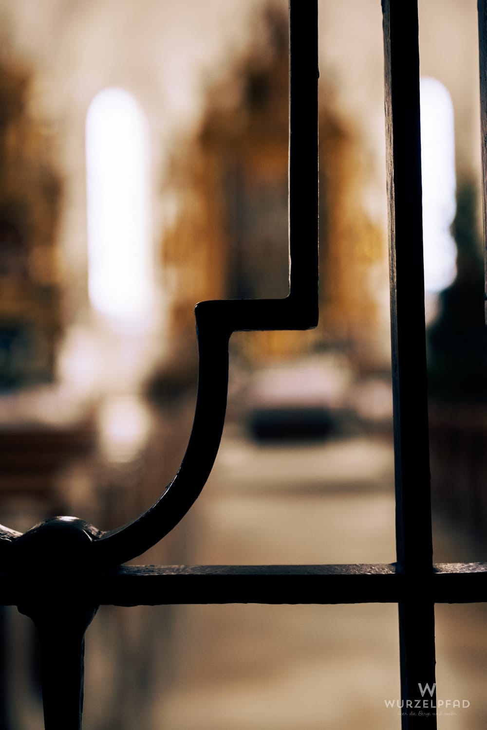 Detail eines dunklen, L-förmigen Metallgitters im Vordergrund, das den Blick auf den unscharfen, hellen Innenraum einer Kirche freigibt.