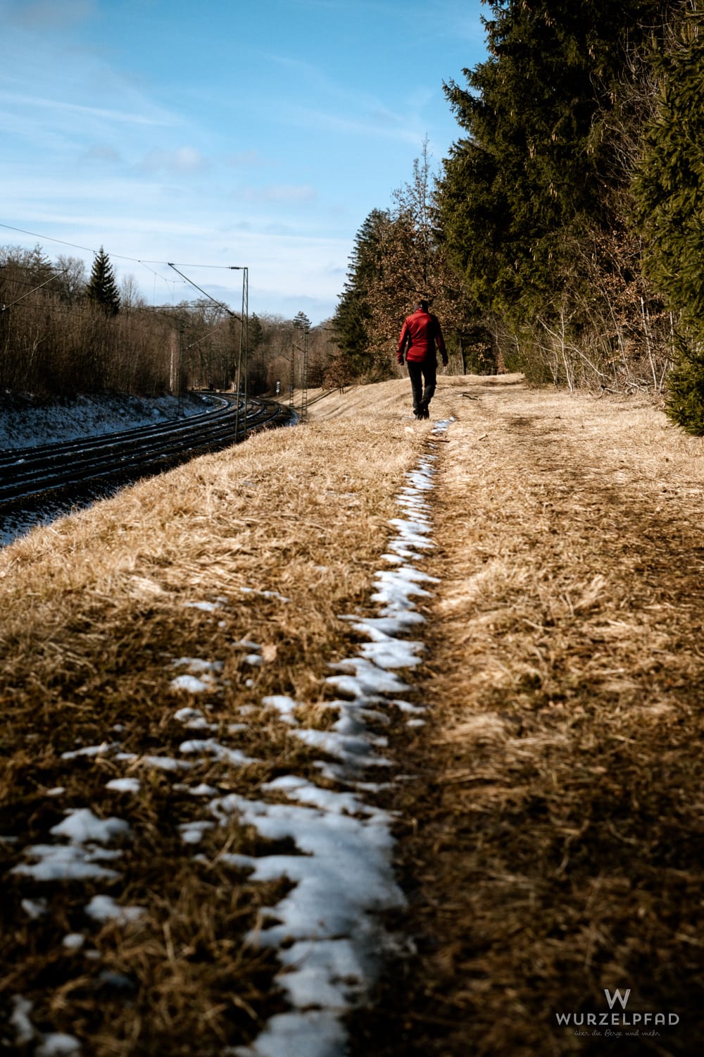Eine Wandersfrau geht auf einem Pfad mit trockenem Gras und Schneeresten entlang von S-Bahn-Gleisen und Bäumen unter blauem Himmel.