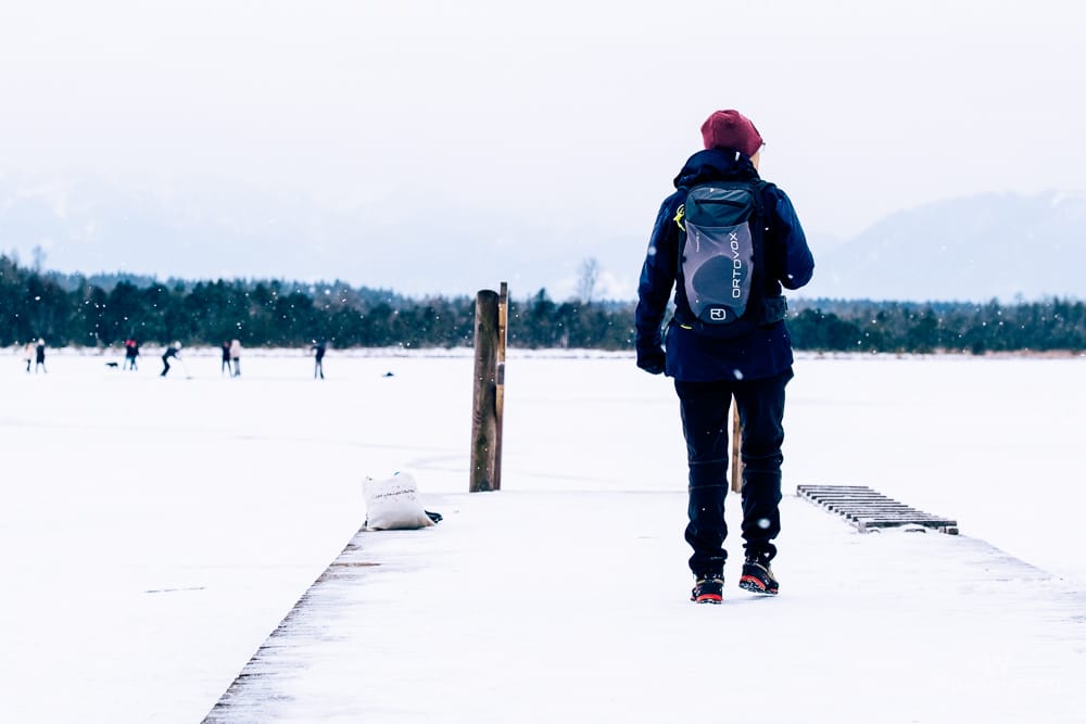 Eine Person mit Rucksack auf einem schneebedeckten Steg am Kirchsee. Im Hintergrund sind Menschen beim Winterspaß auf dem zugefrorenen See und verschneite Berge zu sehen. Es schneit leicht.