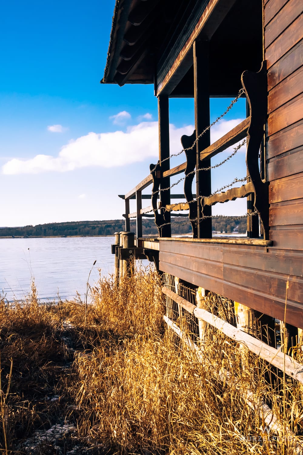 Ein hölzernes Bootshaus am Ufer des Ammersees, dessen Geländer mit Stacheldraht umwickelt ist. Im Vordergrund trockenes Schilf, dahinter der teilweise gefrorene See unter blauem Himmel.