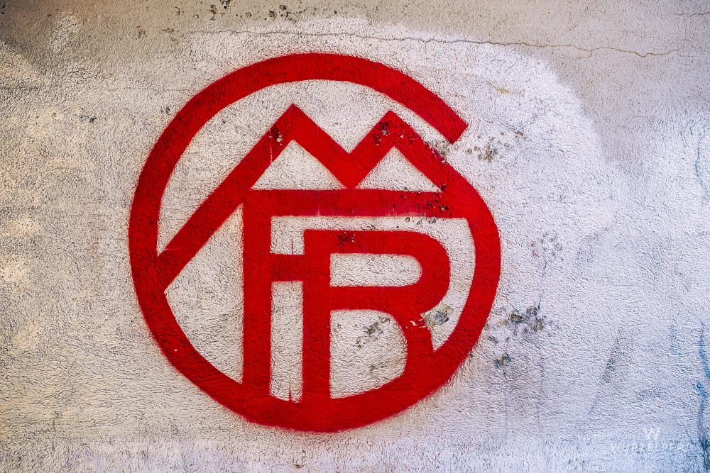 Rotes Graffiti des alten FC Bayern München Wappens auf einer rauen, hellen Wand. Das Logo zeigt ein M über FB in einem Kreis.