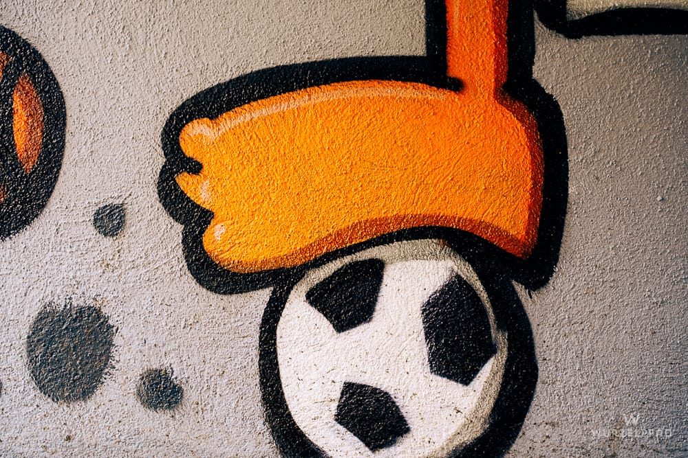 Graffiti auf einer Wand: Ein orangefarbener Fuß tritt einen schwarz-weißen Fußball. Links sind schwarze Punkte zu sehen.