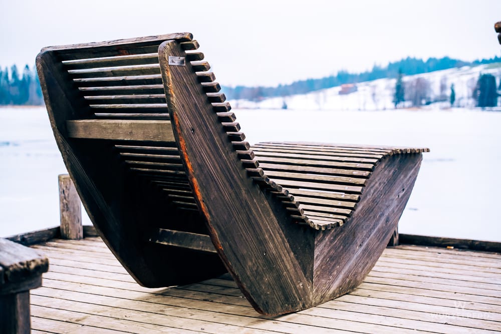 Eine dunkle Holzliege, als "Naturkino-Sitzbank" in erster Reihe auf einem Holzsteg, blickt auf einen zugefrorenen See und eine schneebedeckte Winterlandschaft.