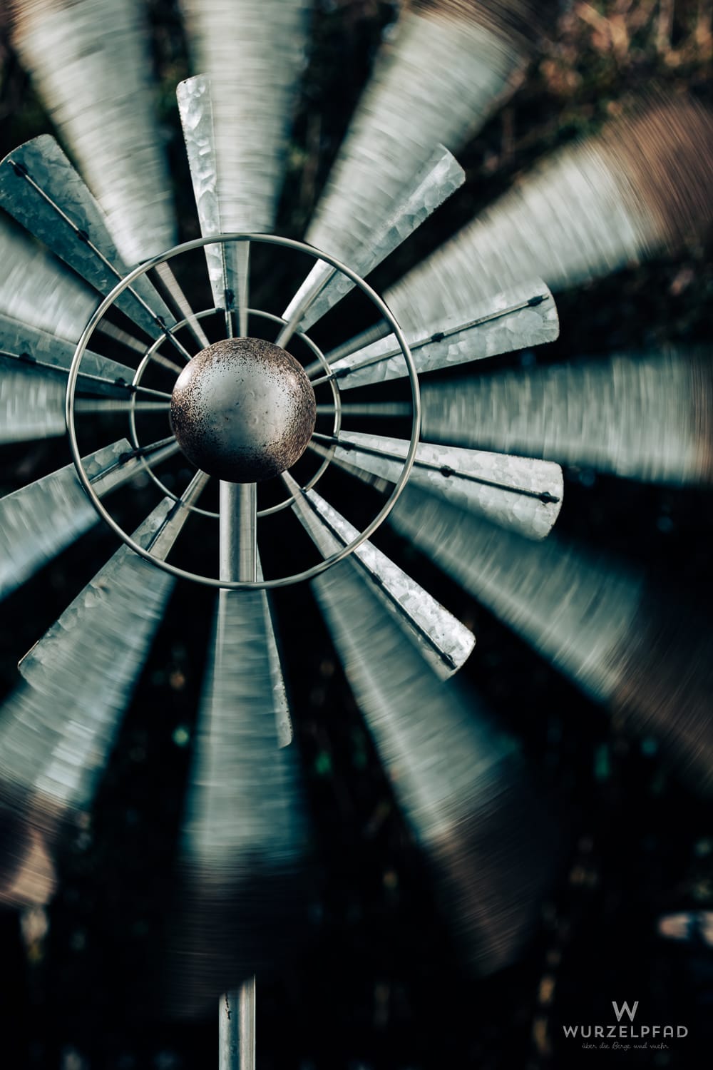 Nahaufnahme eines sich schnell drehenden Windrads aus Metall im Garten. Die Flügel sind durch Bewegungsunschärfe verwischt, die zentrale Kugel ist leicht rostig.