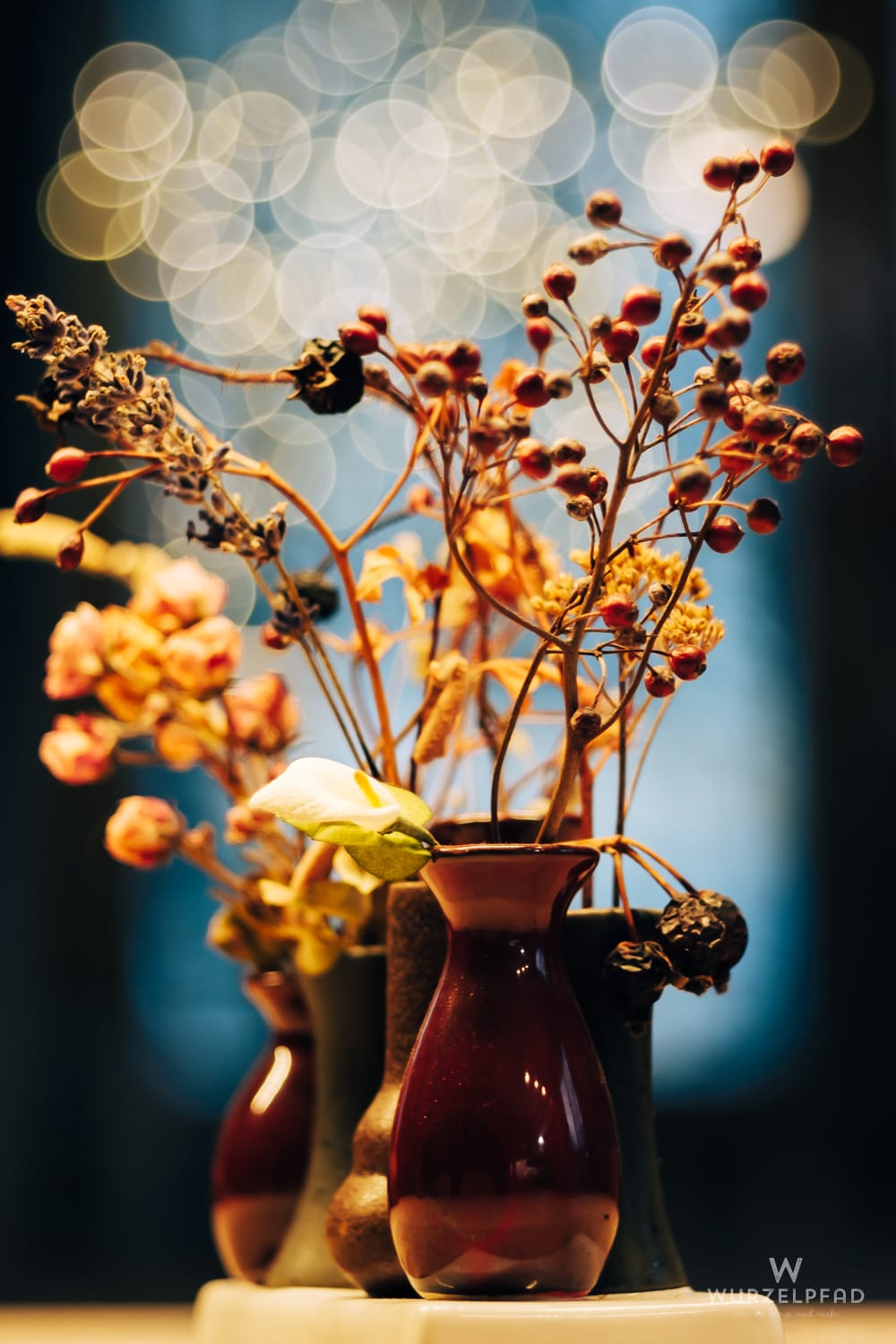 Nahaufnahme einer Blumendeko mit getrockneten roten Beeren, Ästen und einer weißen Calla in dunklen Vasen. Im Hintergrund viele helle, unscharfe Bokeh-Lichtkreise.