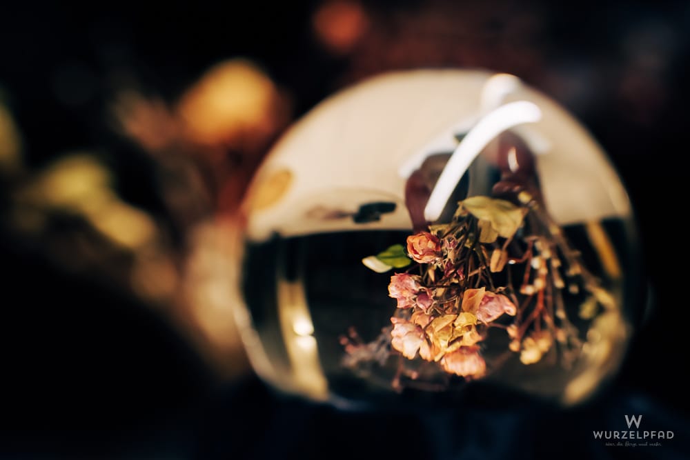 Ein Blumengesteck aus getrockneten, verblühten Rosen in einer Glaskugel, mit unscharfem, dunklem Hintergrund.