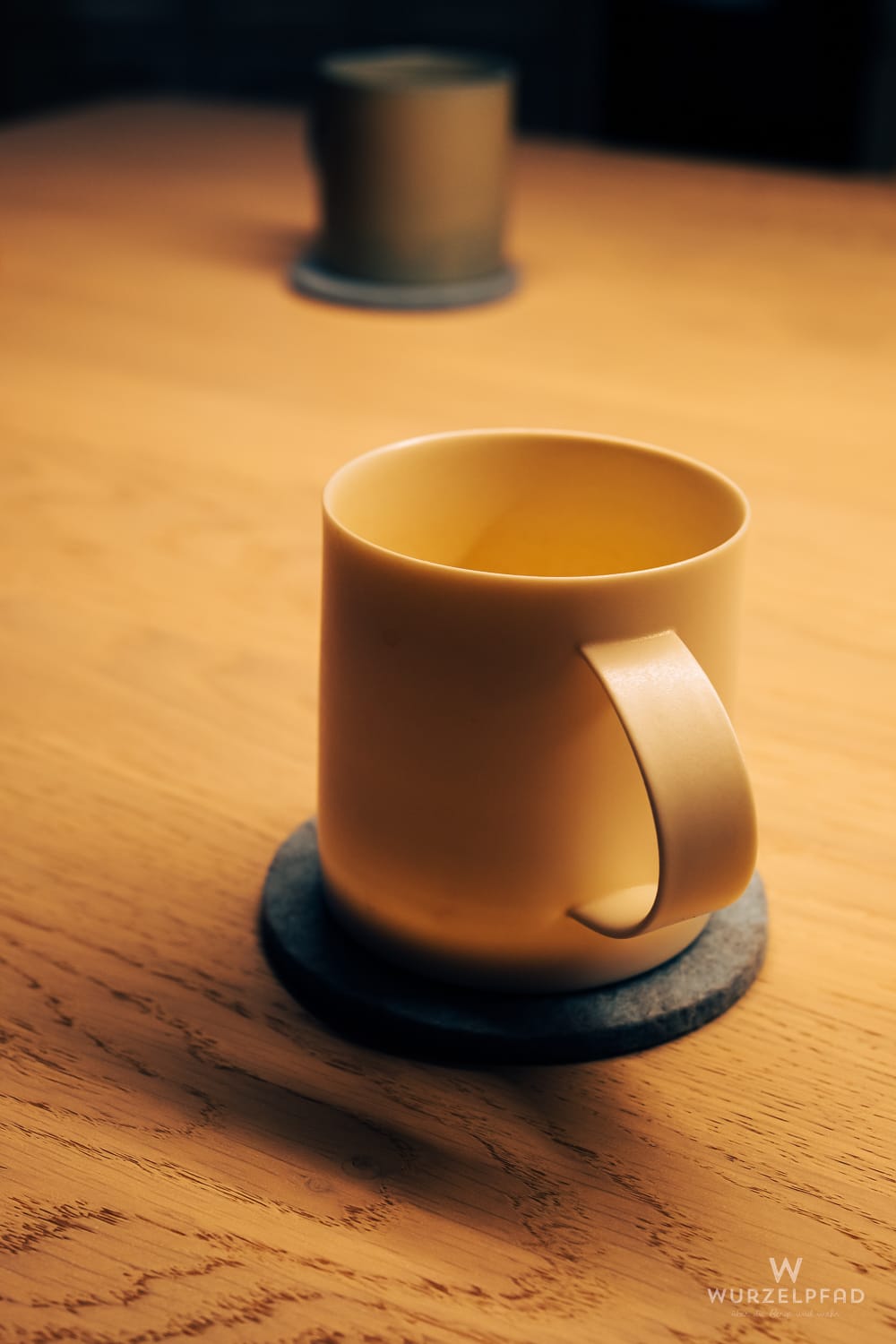 Eine beige Tasse mit Henkel steht auf einem grauen Untersetzer auf einem Holztisch. Eine zweite Tasse ist unscharf im Hintergrund. Warmes Licht erzeugt Schatten.