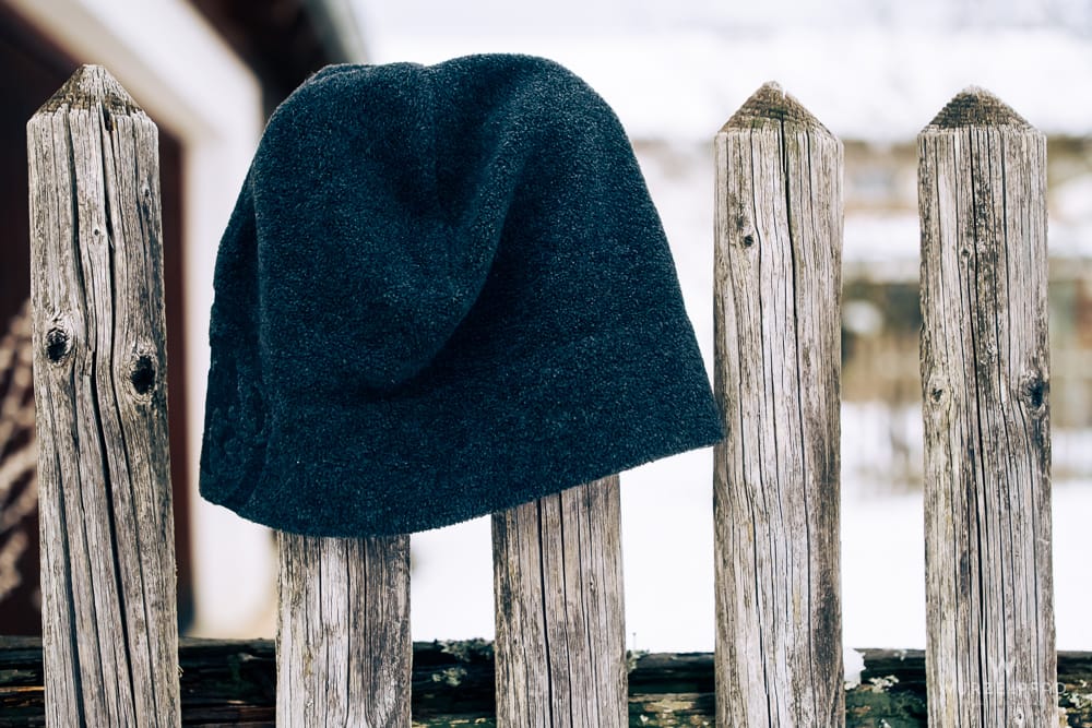 Eine dunkelblaue Strickmütze hängt auf einem verwitterten Holzpfosten eines Zauns. Im Hintergrund ist eine unscharfe, schneebedeckte Landschaft zu sehen.