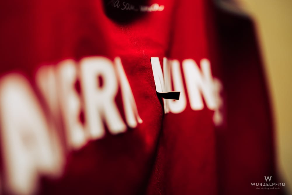 Rotes Fußballtrikot mit unscharfem weißen Schriftzug "BAYERN MÜNCHEN. Der Buchstabe "M" löst sich vom Trikot ab.