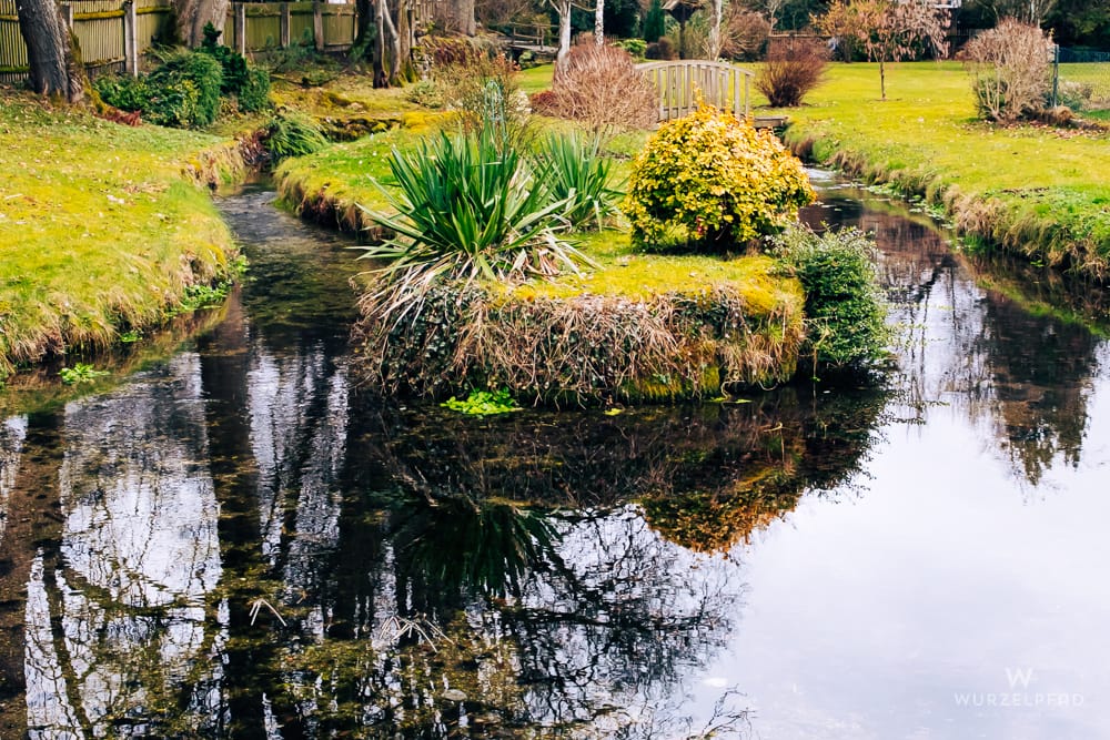Gartenansicht mit einem Bach, der eine Insel mit grünen Pflanzen und einem gelben Busch umfließt. Im Hintergrund eine Holzbrücke. Bäume spiegeln sich im Wasser.