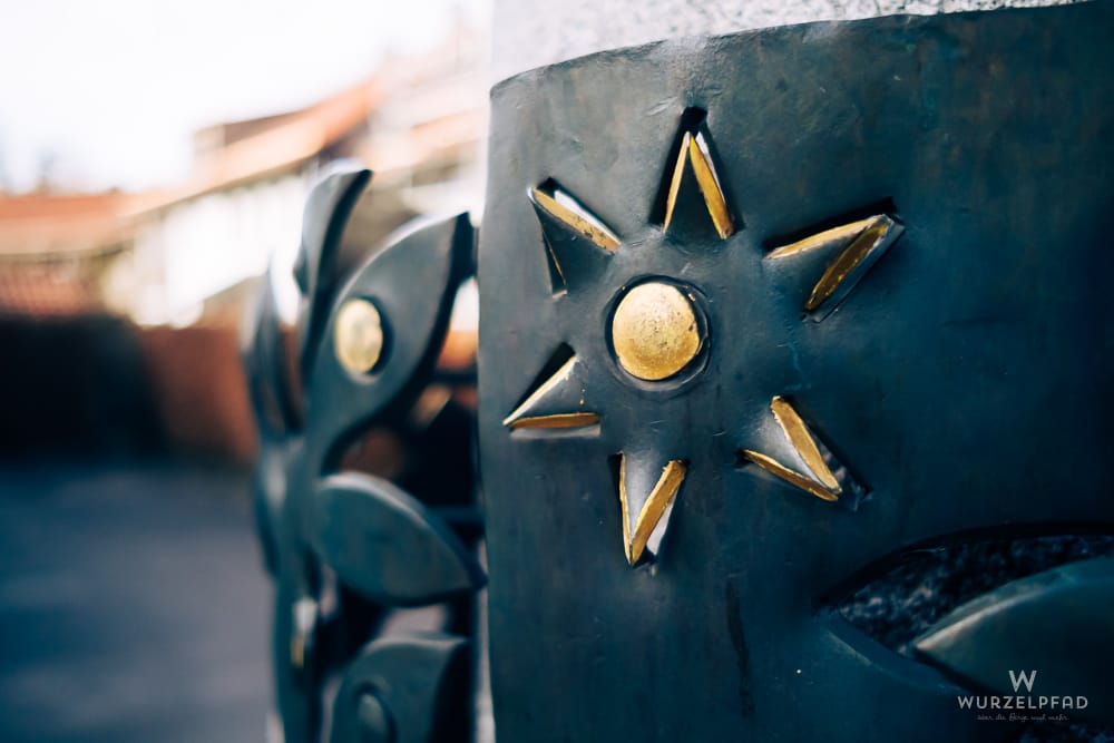 Detailaufnahme einer dunklen Metallskulptur mit goldenen Stern- und Blattmotiven, die Teil eines Brunnens ist.