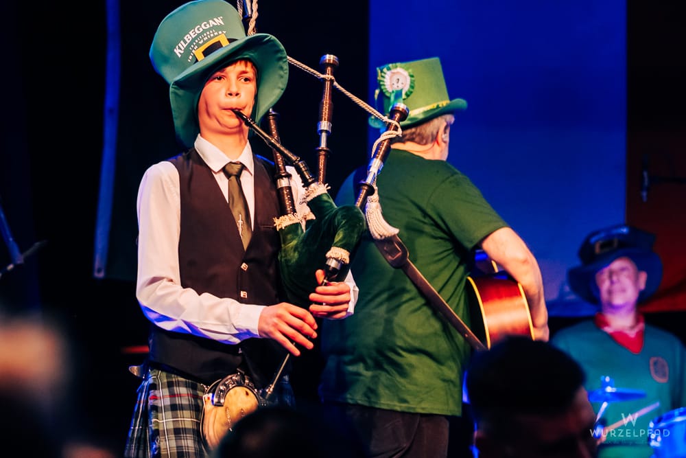 Junge Person in Kilts und grüner Kappe spielt Dudelsack, während ein Mann Gitarre spielt und ein anderer Schlagzeug spielt.