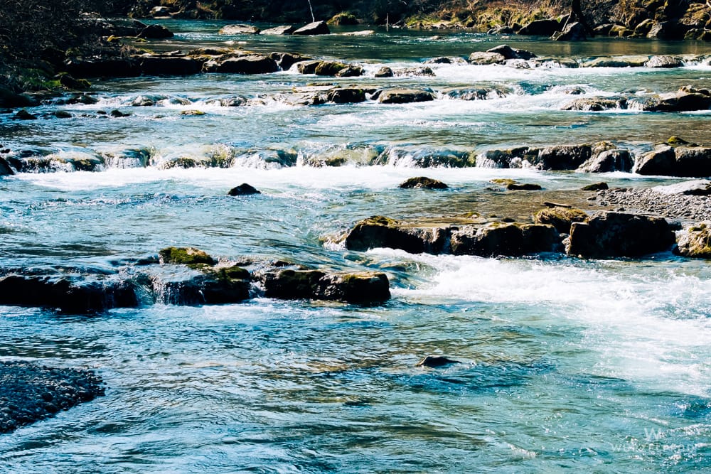 Ein Fluss mit klarem, türkisfarbenem Wasser fließt über Felsen und kleine Kaskaden. Im Hintergrund ist ein dunkler Wald zu sehen.