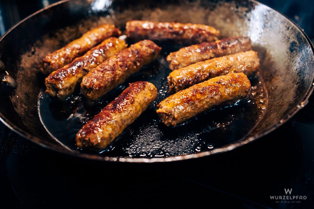 Mehrere Cevapcici brutzeln in einer schwarzen Eisenpfanne in heißem Öl.